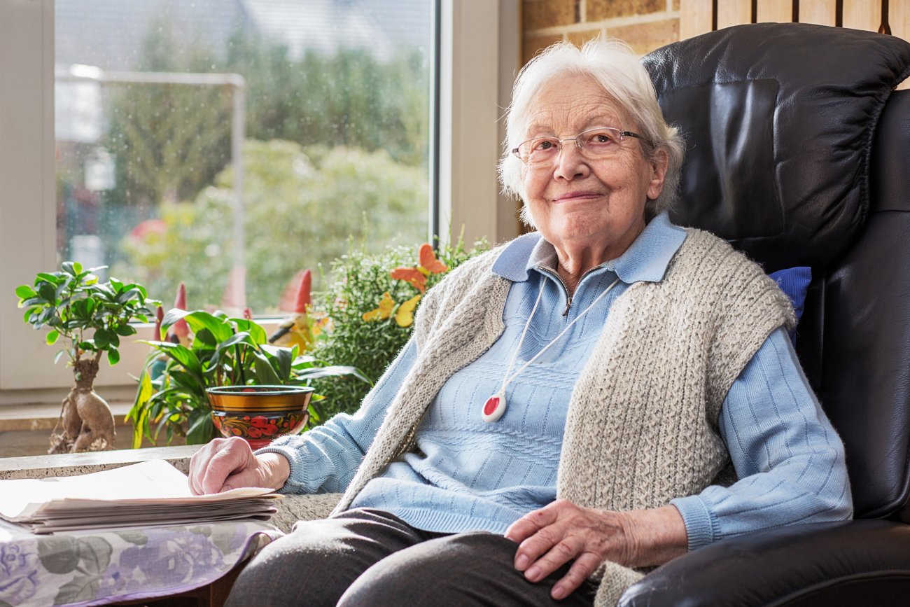 Une femme âgée est assise dans un fauteuil et porte le bouton de téléalarme autour du cou.