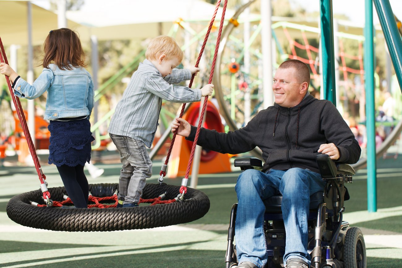 Un homme dans un fauteuil roulant joue avec 2 petits enfants valides dans un parc de jeux