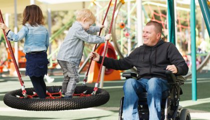 Un homme dans un fauteuil roulant joue avec 2 petits enfants valides dans un parc de jeux